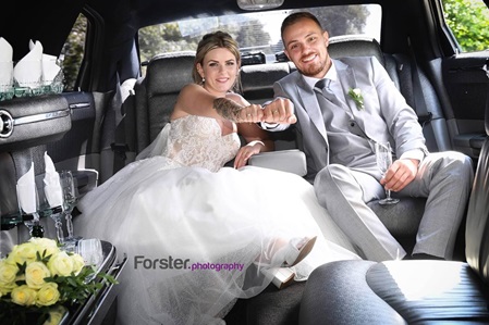 Brautpaar sitzt beim Fotoshooting lächelnd in einer Limousine und halten ihre Trauringe in die Kamera