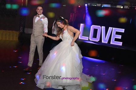 Brautpaar tanzt auf seiner Hochzeitsfeier ausgelassen vor einem Love-Schild mit vielen bunten Lichtern