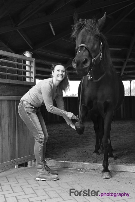 Junge Frau lächelt und kratzt den Huf eines schwarzen Pferdes bei einem Tier-Fotoshooting in Iserlohn.