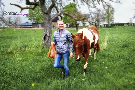 Junge blonde Frau führt lächelnd ein braunes Pferd auf einer grünen Koppel bei einem Fotoshooting in Iserlohn.
