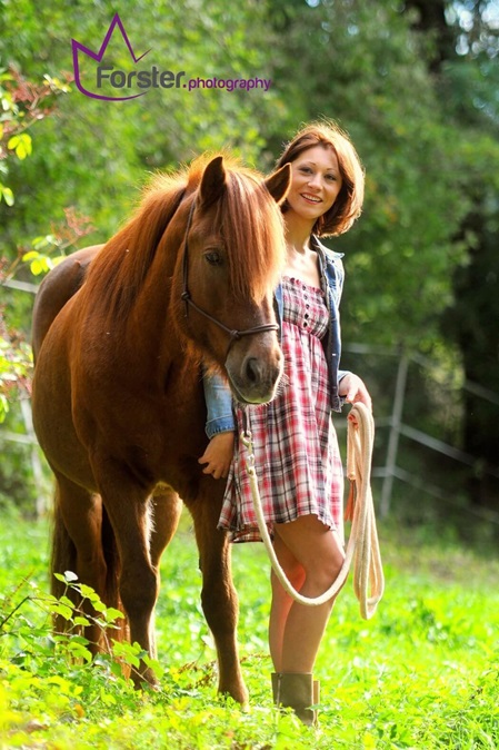 Junge Frau im Sommerkleid mit braunem Pferd auf einer grünen Wiese.
