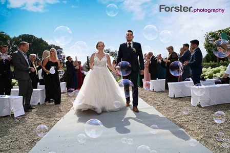 Ein Brautpaar im Hochzeits-Outfit geht nach der Trauung Hand in Hand an den Gästen mit Seifenblasen vorbei
