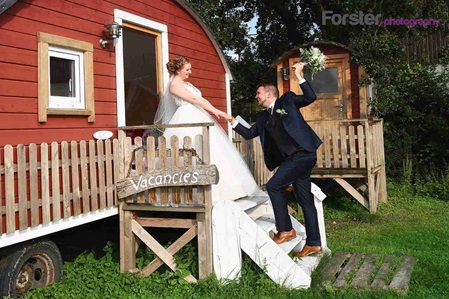 Ein Brautpaar im Hochzeits-Outfit posiert beim Fotoshooting vor einem roten Tiny-House, er hält die Hand der Braut