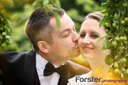 Ein Brautpaar im Hochzeits-Outfit steht beim Fotoshooting dicht zusammen, er küsst die Braut