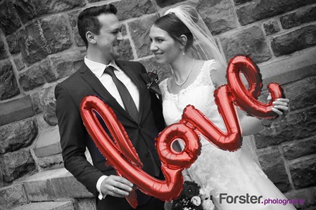 Ein Brautpaar im Hochzeits-Outfit beim Fotoshooting schaut sich mit LOVE-Luftballon verliebt und glücklich an