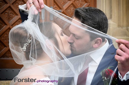 Ein Brautpaar im Hochzeits-Outfit steht beim Fotoshooting dicht zusammen, er hält den Schleier und küsst die Braut
