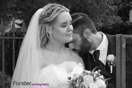 Ein Brautpaar im Hochzeits-Outfit steht beim Fotoshooting dicht zusammen, er küsst die Braut