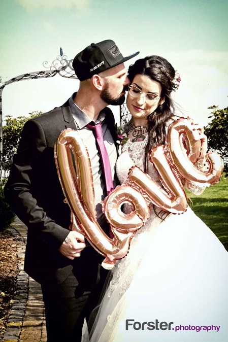 Ein Brautpaar im Hochzeits-Outfit posiert beim Fotoshooting in einem Park mit einem Love-Luftballon in der Hand
