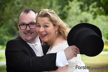 Elegantes Brautpaar lacht beim Hochzeits-Fotoshooting in die Kamera, der Bräutigam hat seinen Zylinder in der Hand