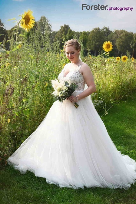 Eine Braut im weißen Hochzeitskleid steht vor einem Feld mit Sonnenblumen und blickt auf den Brautstrauß hinab