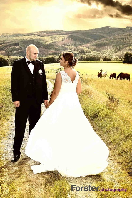 Brautpaar im Hochzeits-Outfit steht beim Fotoshooting lächelnd zusammen auf einem Feld vor der untergehenden Sonne