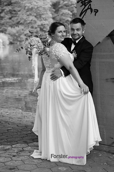 Ein Brautpaar im Hochzeits-Outfit umarmt sich und posiert beim Fotoshooting vor dem Seilersee und lächelt in die Kamera