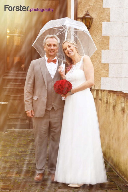 Brautpaar im Hochzeits-Outfit steht beim Fotoshooting unter einem Regenschirm lachend vor der Kamera