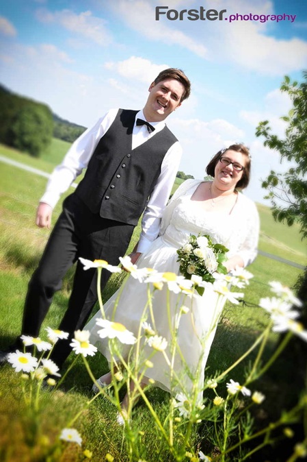 Ein Brautpaar im Hochzeits-Outfit posiert beim Fotoshooting vor einem Feld und lächelt in die Kamera