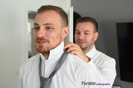 Ein Bräutigam steht vor der Hochzeit beim getting ready Fotoshooting im Wohnzimmer, Trauzeuge knotet die Krawatte