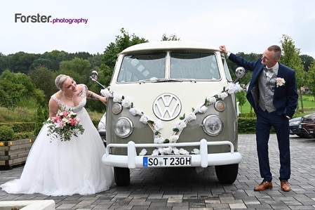 Hochzeitspaar steht beim Fotoshooting jeweils links und rechts von einem VW-Bulli und schauen sich dabei an