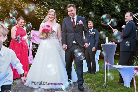 Ein Brautpaar im Hochzeits-Outfit geht nach der Trauung Hand in Hand an den Gästen mit Seifenblasen vorbei