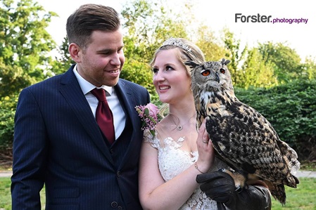 Ein Brautpaar im Hochzeits-Outfit steht beim Fotoshooting nebeneinander, die Braut hat eine große Eule auf dem Arm