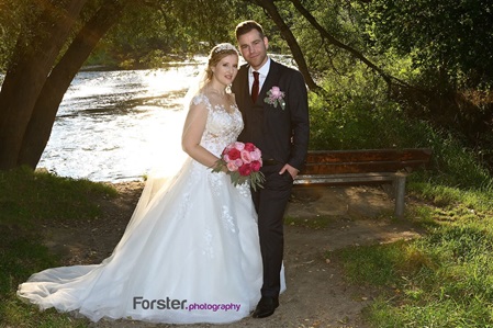 Eine Braut im weißen Hochzeitskleid steht beim Fotoshooting mit dem Bräutigam am Fluss und lächelt in die Kamera