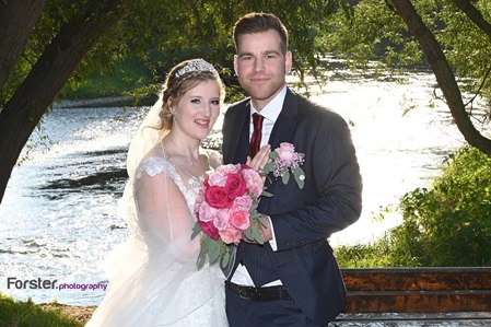 Eine Braut im weißen Hochzeitskleid steht beim Fotoshooting mit dem Bräutigam am Fluss und lächelt in die Kamera