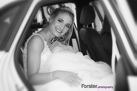 Eine Braut im weißen Hochzeitskleid sitzt lächelnd im Brautauto und steigt aus.