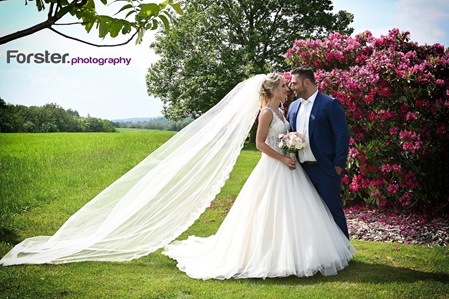 Eine Braut im weißen Hochzeitskleid mit langem Schleier steht mit dem Bräutigam beim Fotoshooting in einem Park