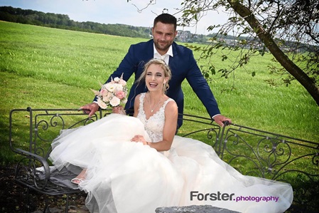 Eine Braut im weißen Hochzeitskleid sitzt beim Fotoshooting und lächelt mit ihrem Bräutigam in die Kamera