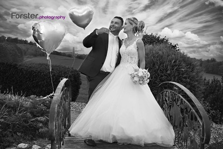 Ein Brautpaar im Hochzeits-Outfit steht beim Fotoshooting auf einer Brücke im Park vor einem dramatischen Himmel