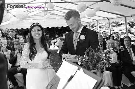 Ein Brautpaar im Hochzeits-Outfit steht bei der freien Trauung vor seinen Gästen und schaut lachend in die Kamera
