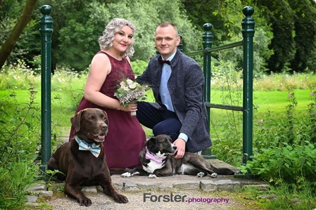 Brautpaar im Hochzeits-Outfit beim Fotoshooting hockt auf einer Brücke mit zwei Hunden und schauen in die Kamera