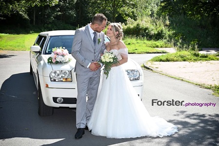 Ein Brautpaar im Hochzeits-Outfit steht beim Fotoshooting vor einer Limousine in einem Park und schaut sich an