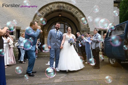 Ein Brautpaar im Hochzeits-Outfit tritt glücklich aus der Kirche. Die Gäste empfangen sie mit Seifenblasen