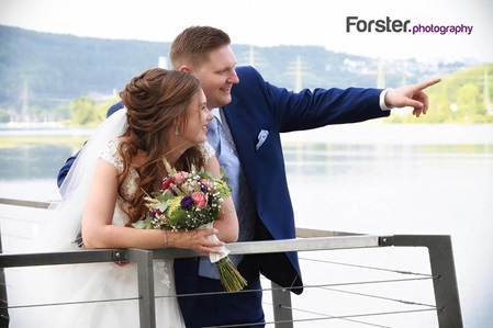 Ein Brautpaar im Hochzeits-Outfit steht beim Fotoshooting am See. Der Bräutigam zeigt mit der Hand in die Ferne