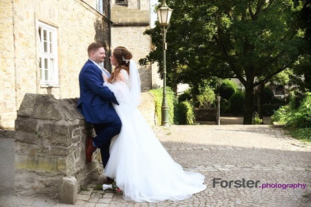 Eine Braut im Hochzeitskleid mit langem Schleier steht beim Fotoshooting mit ihrem Bräutigam an einer Steinmauer