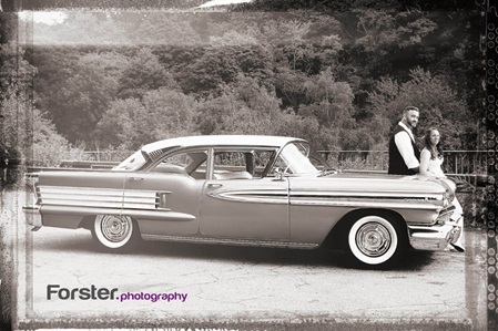 Ein Brautpaar im Hochzeits-Outfit sitzt beim Fotoshooting vorne auf einem Oldtimer