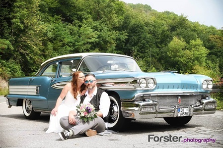 Ein Brautpaar im Hochzeits-Outfit sitzt beim Fotoshooting vor einem Oldtimer. Die Braut küsst den Bräutigam zärtlich