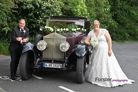 Ein Brautpaar im Hochzeits-Outfit beim Fotoshooting steht neben einem Oldtimer und lächeln glücklich in die Kamera