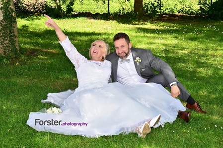 Brautpaar sitzt beim Fotoshooting auf einer Wiese. Das weiße Brautkleid ist ausgebreitet. Braut zeigt in die Höhe