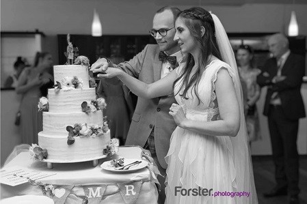 Brautpaar beim Fotoshooting auf der Hochzeitsfeier. Sie schneiden die Hochzeitstorte an. Gäste im Hintergrund