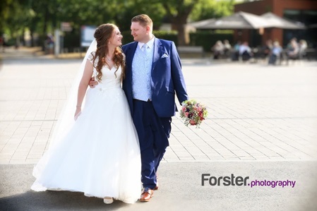 Brautpaar am Hochzeitstag beim Fotoshooting auf einem Platz schauen sich verliebt an, Ehemann hält den Brautstrauß