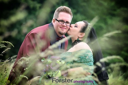 Brautpaar steht beim Fotoshooting auf einer Wiese, die Braut küsst den Ehemann auf die Wange.