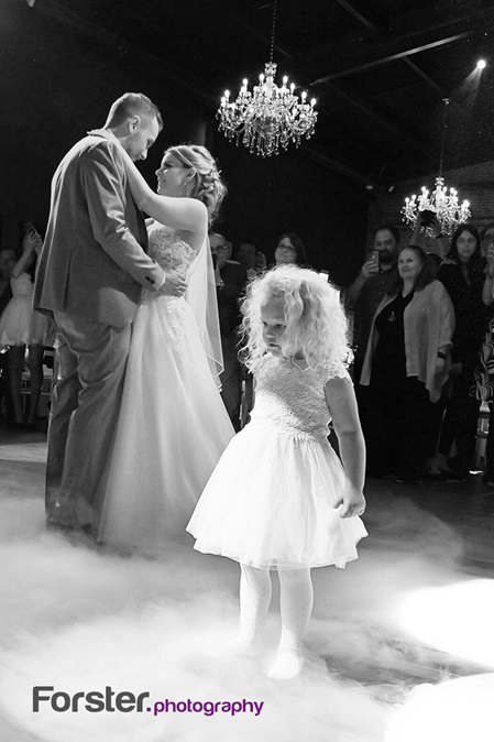 Hochzeitspaar tanzt verliebt mit Bodennebel den ersten Tanz. Vorne steht ein kleines Mädchen im weißen Kleid