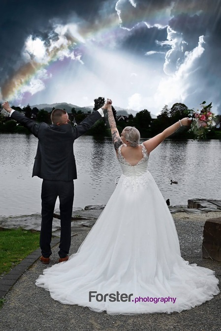 Brautpaar beim Fotoshooting stehen am See mit Regenbogen. Sie stecken die Arme in die Höhe und schauen sich an