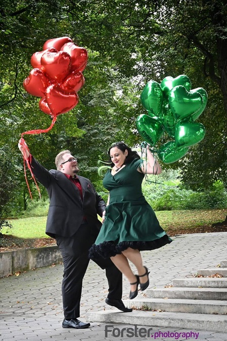 Ein elegantes Brautpaar beim Fotoshooting mit roten und grünen Luftballons. Die Braut springt von einer Treppe