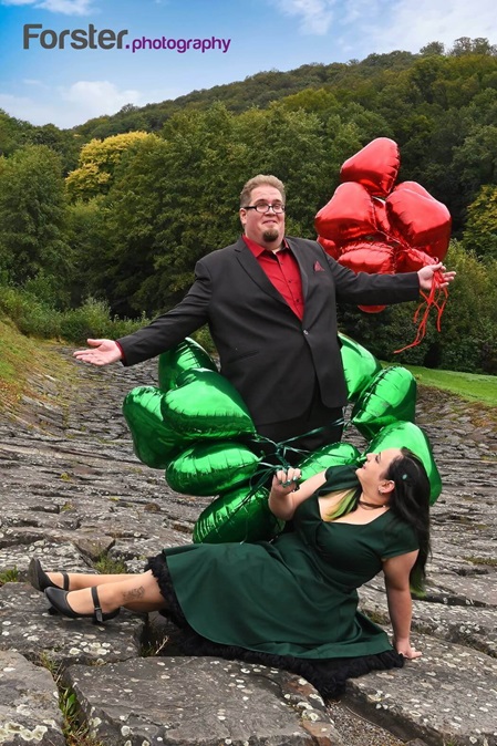 Ein elegantes Brautpaar beim Fotoshooting mit roten und grünen Luftballons. Die Braut sitzt zu Füßen des Bräutigams