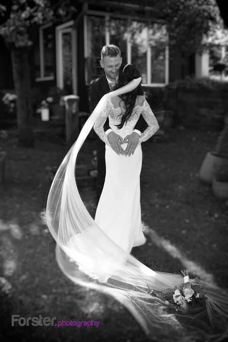 Eine Braut im Hochzeitskleid mit langem Schleier beim Fotoshooting im Garten. Sie formen mit ihren Händen ein Herz.
