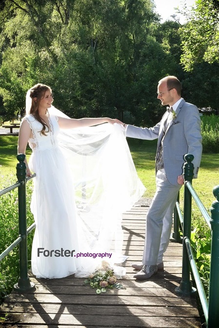 Eine Braut im Hochzeitskleid steht beim Fotoshooting mit ihrem Bräutigam auf einer Brücke und reichen sich die Hand
