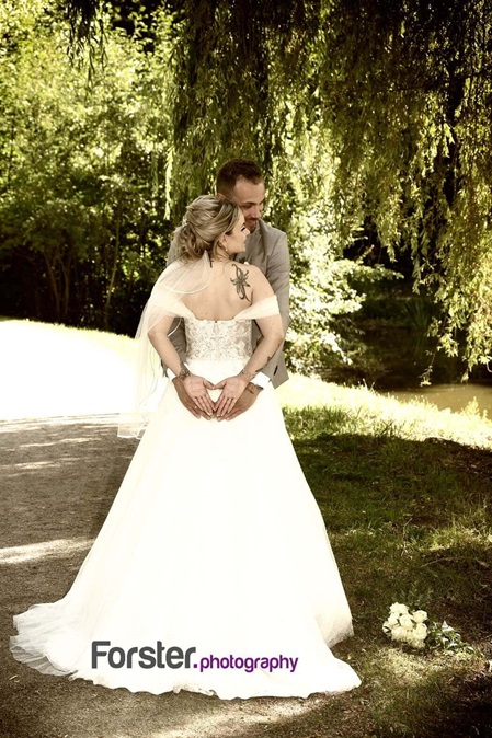 Eine Braut im weißen Hochzeitskleid beim Fotoshooting im Park. Sie formt mit dem Bräutigam mit ihren Händen ein Herz